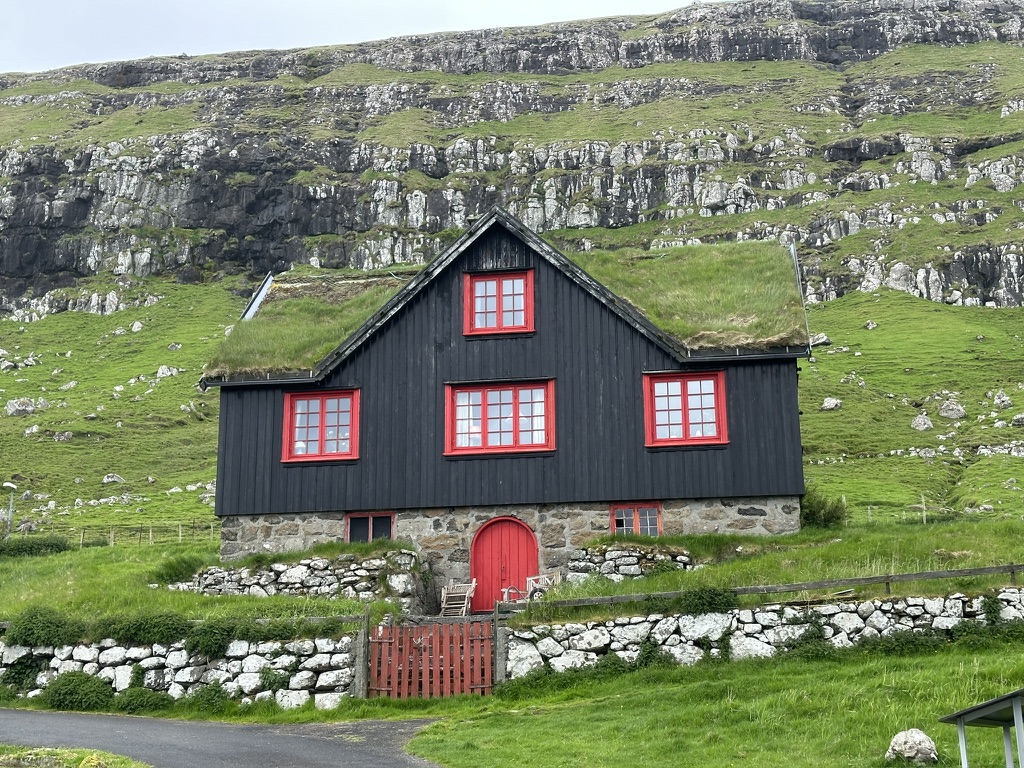 Schwarzes Haus mit Grasdach und roten Fenstern vor grünen Hängen auf den Färöern